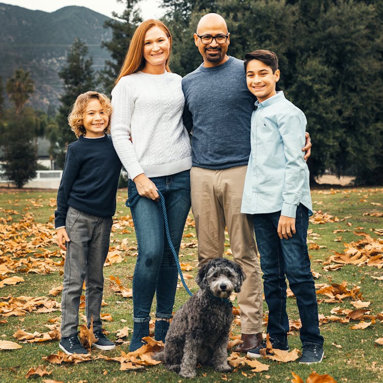 A family of five with two boys and a dog standing on a lawn covered with autumn leaves.