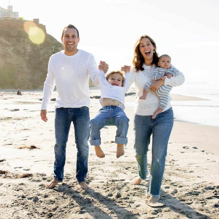 A family of four walking on a beach, smiling, with two parents holding their children's hand.