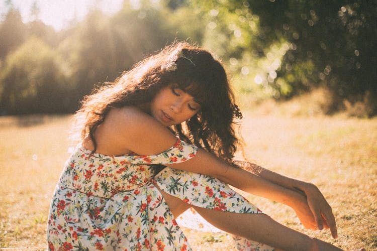 A woman in a floral dress sitting pensively in a sunlit field