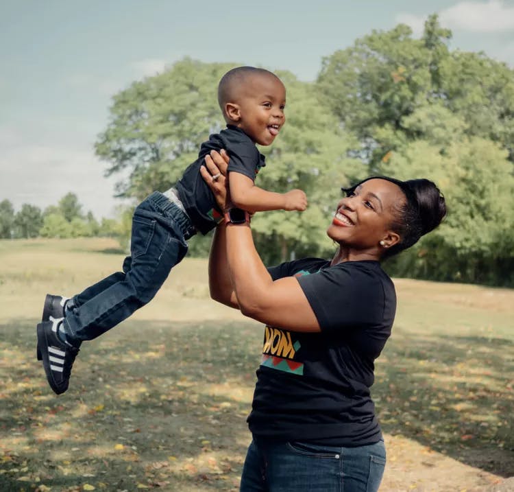 A woman happily lifting a young boy in the air at a park.