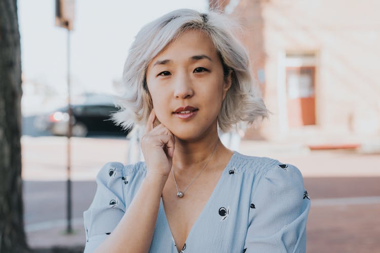 A portrait of a woman with short silver hair wearing a light blue dress and touching her chin