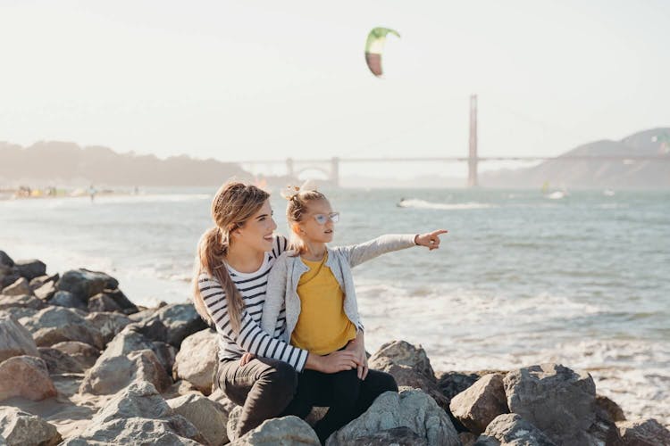 A woman and a young girl sitting on rocks by the sea, the child pointing towards the water