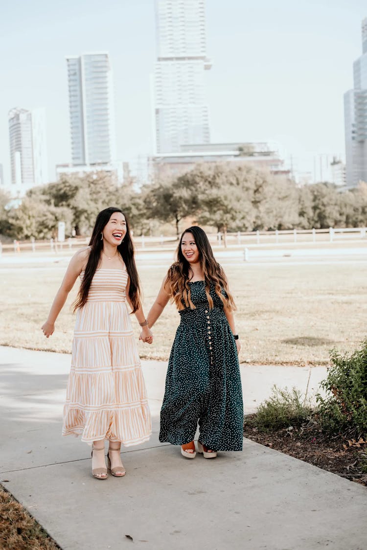 Two women walking and smiling in a park with city buildings in the background.