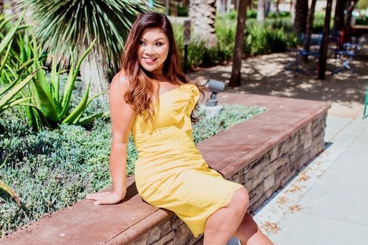 A woman in a yellow dress sitting on a stone bench with greenery around her.